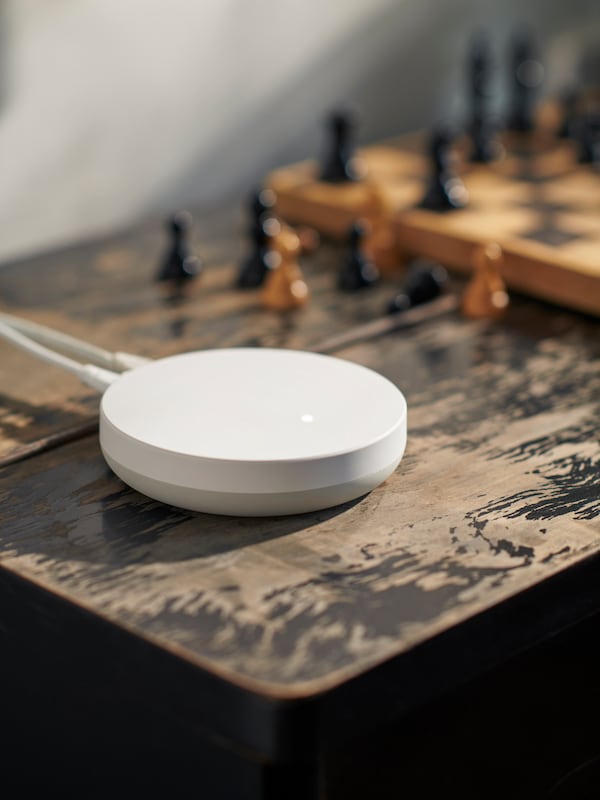 A white, round DIRIGERA hub for smart products is placed on an old wooden table next to a game of chess.