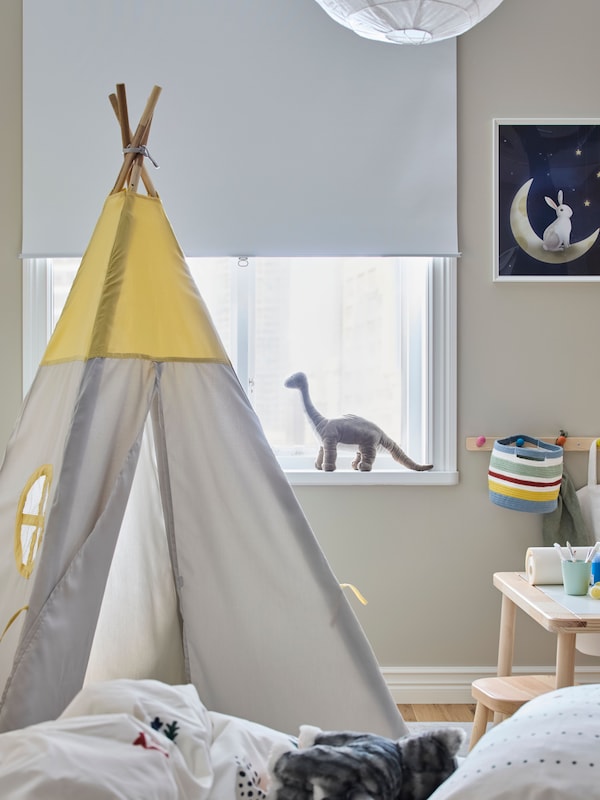 A white TRETUR block-out roller blind in a window with a soft toy dinosaur/Brontosaurus on the sill, plus a children’s tent.