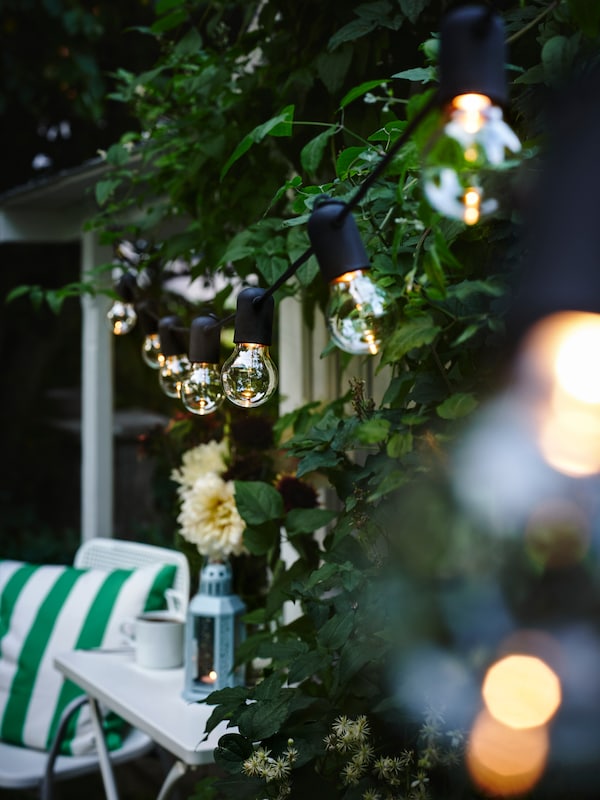 A black SVARTRÅ LED lighting chain with 12 lights is hanging in a tree above a table and a chair with a green-white cushion.
