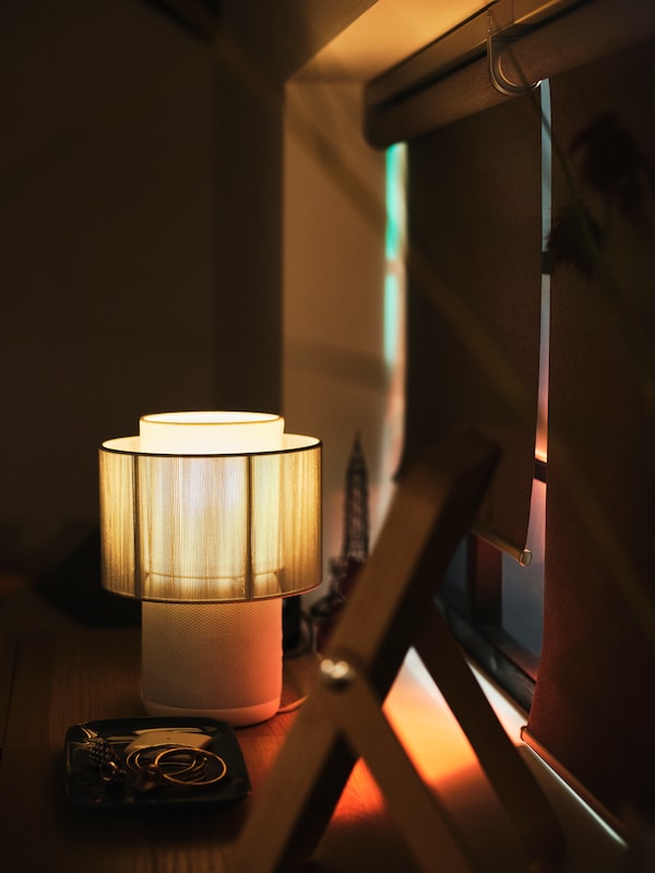 A lit SYMFONISK speaker lamp with Wi-Fi stands beside a window with two FYRTUR block-out roller blinds in a dark room.