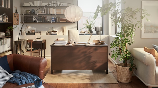 An IKEA IDANÄS desk in brown in the middle of a living room with dark wood floors and white walls, over a beige rug.