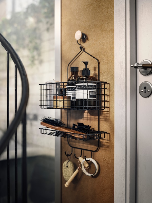 A close-up of BLECKSJÖN shower hanger in black steel, hanging on a bathroom wall, filled with soap bottles and a brush.