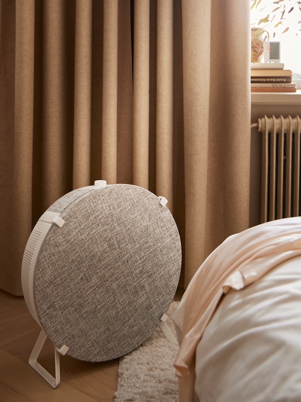 A white STARKVIND air purifier stands at the end of a white KLEPPSTAD bed in front of an ANNAKAJSA room darkening curtain.