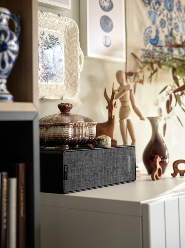 A black SYMFONISK WiFi bookshelf speaker lies on top of a BESTÅ shelf unit with door, along with various decorative objects.