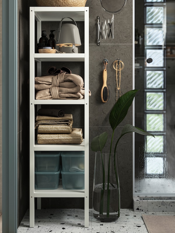 Beige towels, transparent boxes, soap bottles and a wi-fi speaker are stored inside an open MUSKAN shelving unit in white.