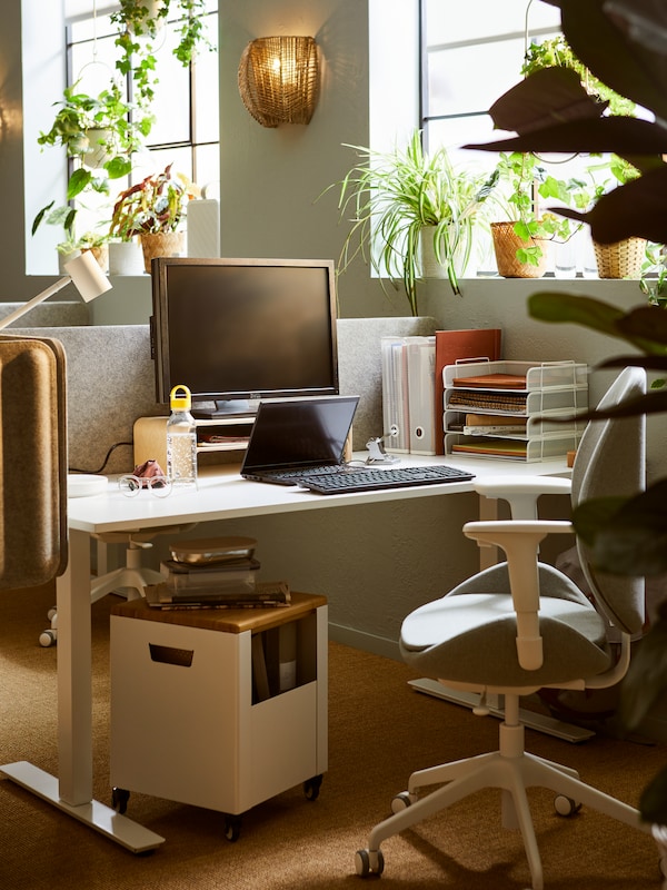 A sunlit workspace with a TROTTEN sit-stand desk and a HATTEFJÄLL office chair, next to a window filled with green plants.
