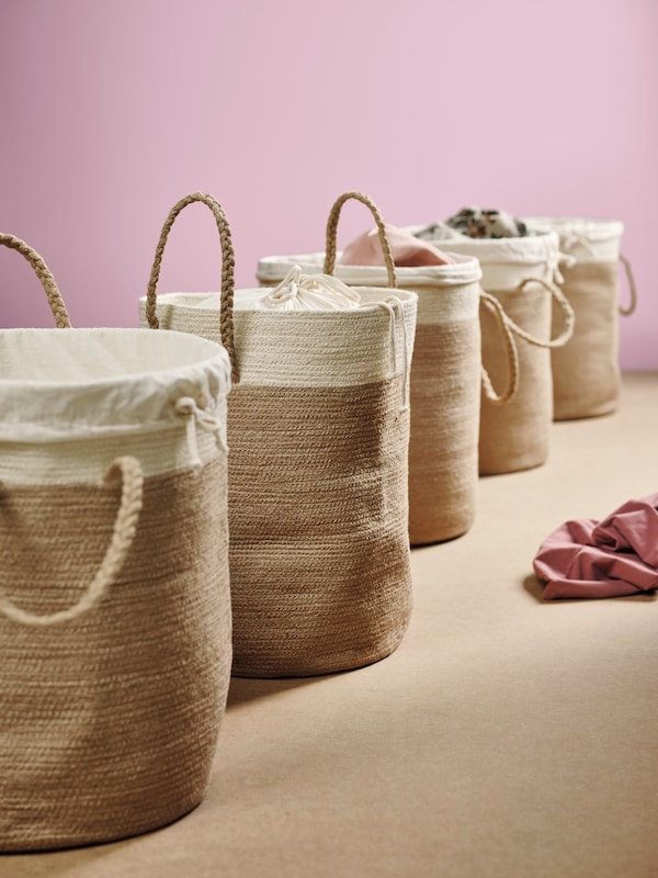 Several LJUNGAN jute laundry bags, some of them with laundry in them, neatly placed in a line against a pink wall.