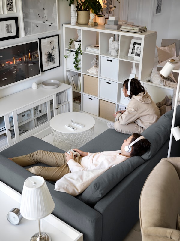 Two people playing video games on an IKEA FRIHETEN corner sofa in dark grey fabric, a white coffee table and tv bench in front, a shelving unit on the side separating the room.