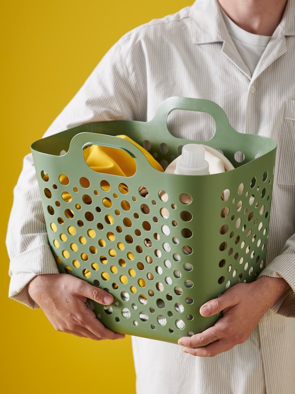 A person, wearing a striped shirt, is holding a green SLIBB flexible laundry basket containing laundry.