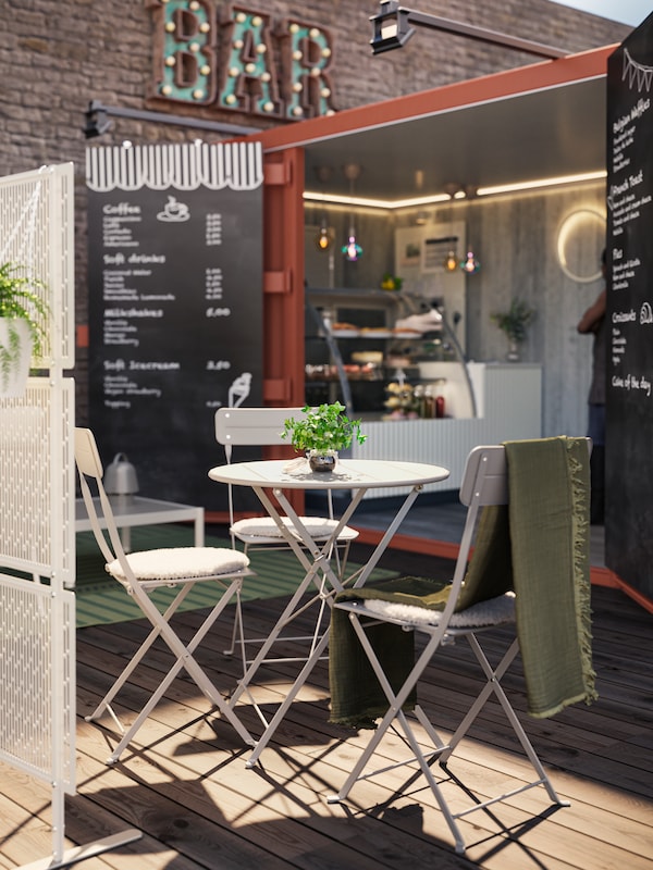 An outdoor lounge area, outside a café, with a grey SUNDSÖ table and two chairs. A green thrown hand on one of the chairs.