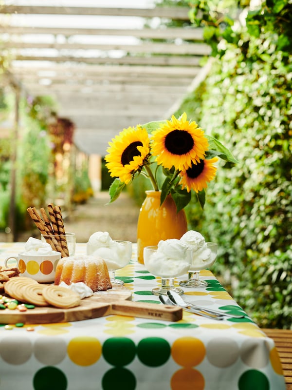A table dressed with a BRÖGGAN tablecloth in white, green and yellow dots, various tableware over it, a yellow vase with sun flowers inside.