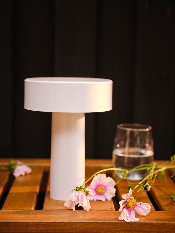 A wooden table on a dark background with a SOLVINDEN LED table lamp, a glass of water and some pink flowers on it.