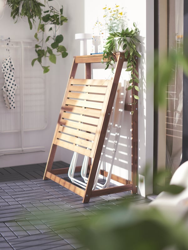 A balcony corner with plants hanging from the wall and a folded, brown, DJUPÖN folding table with two folded chairs inside.