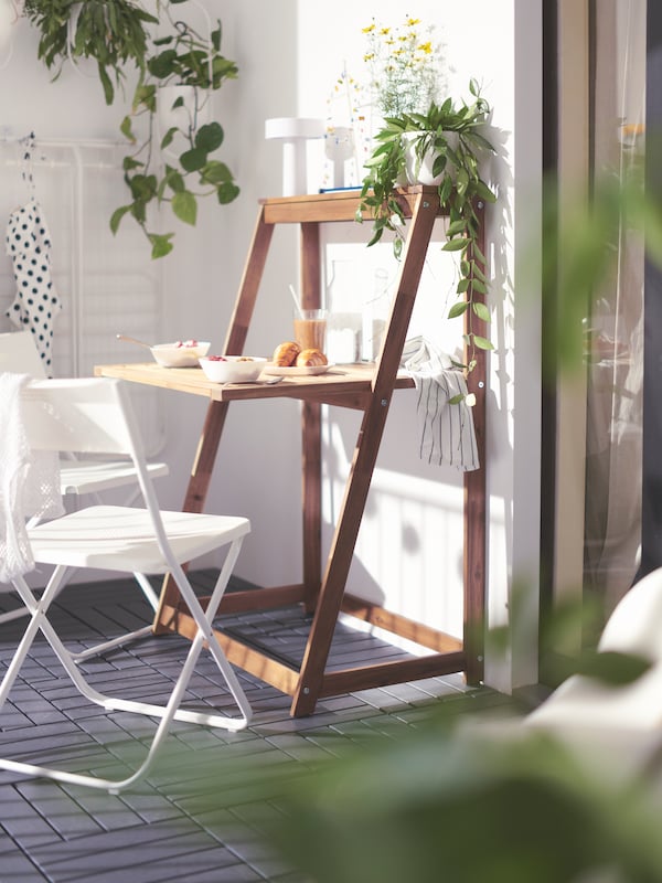 A sunny balcony corner with plants and an opened, brown, DJUPÖN folding table with bowls and glasses, and two white chairs.
