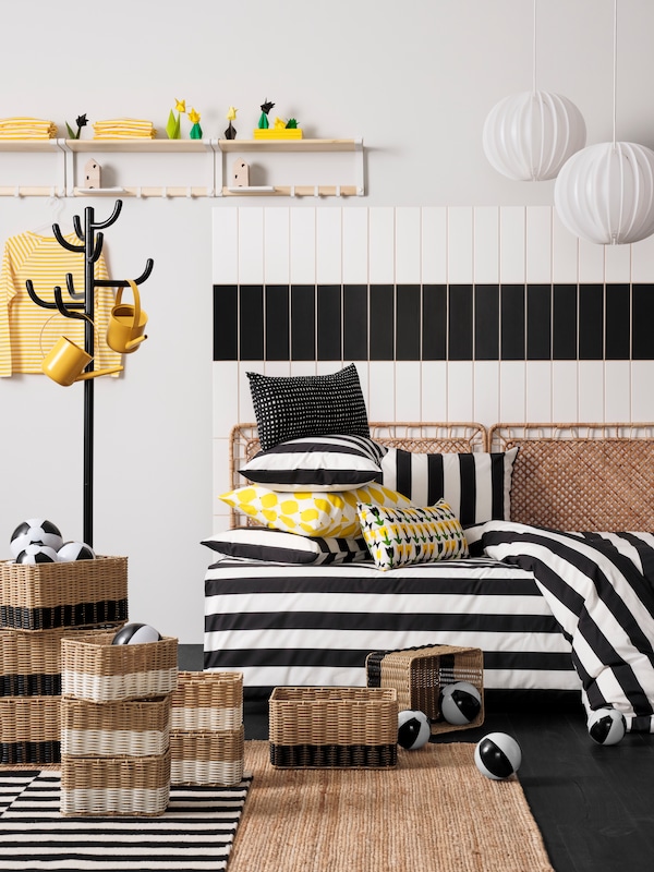 Stacked DJURTRÄNARE baskets next to a white/Tolkning rattan VEVELSTAD bed with black/white/striped SLÅNHÖSTMAL bed linen.