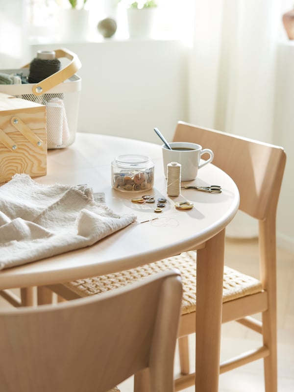 Two SKANSNÄS chairs stand at a SKANSNÄS round extendable table with a RISATORP basket, sewing things and a mug on top of it.