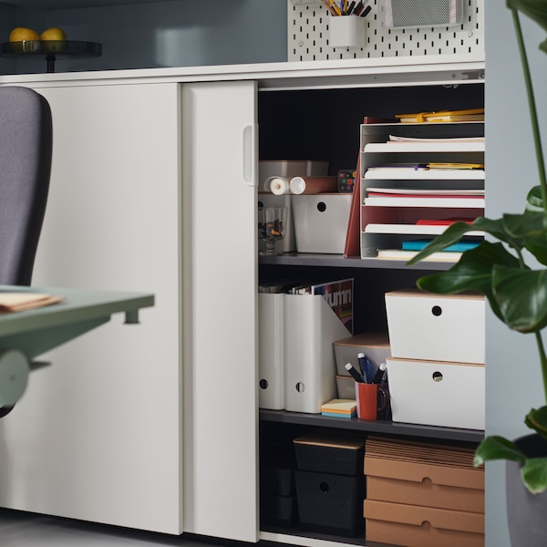 A white GALANT cabinet with sliding doors with one door slightly opened, revealing KUGGIS boxes and office supplies.