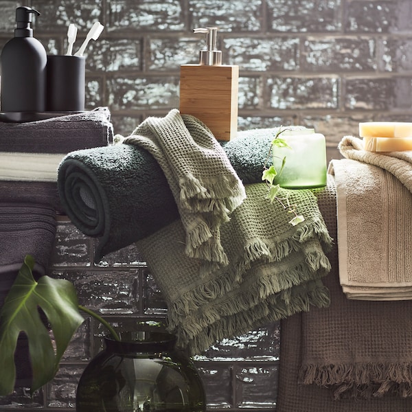 White, light beige, light grey/brown, dark grey and green towels displayed with various accessories in a grey-tiled bathroom.