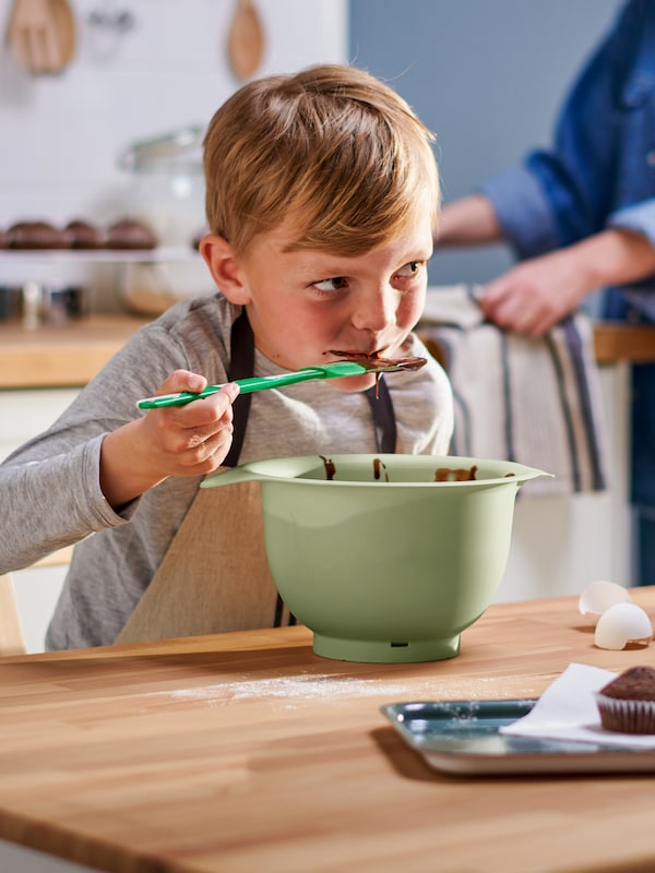 With a green/white GUBBRÖRA spatula, a child in a MARIATHERES apron eats chocolate cake batter from a green mixing bowl.